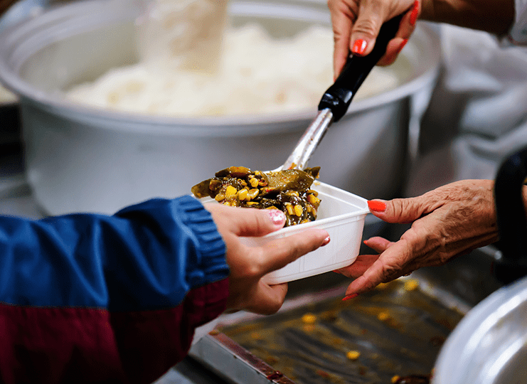 A pair of hands giving out food at a soup kitchen while another hand receives it in a white container.