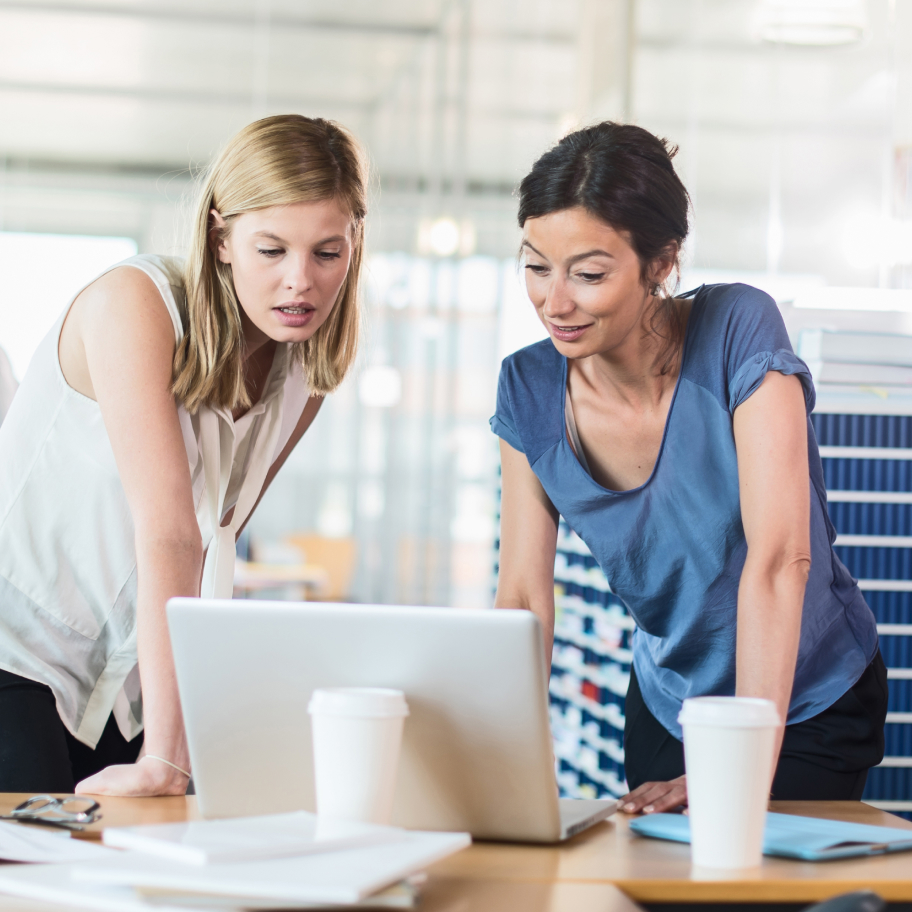 Two people staring at a laptop screen with white coffee cups on a desk.